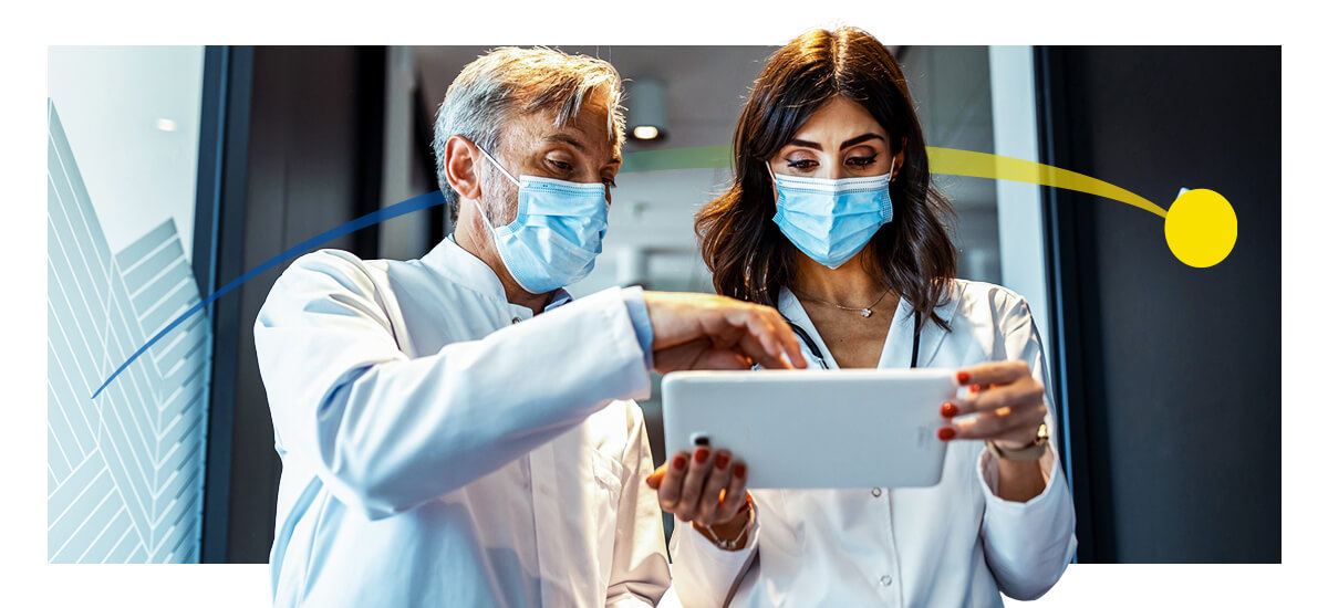 A male and female doctor review a tablet while wearing masks